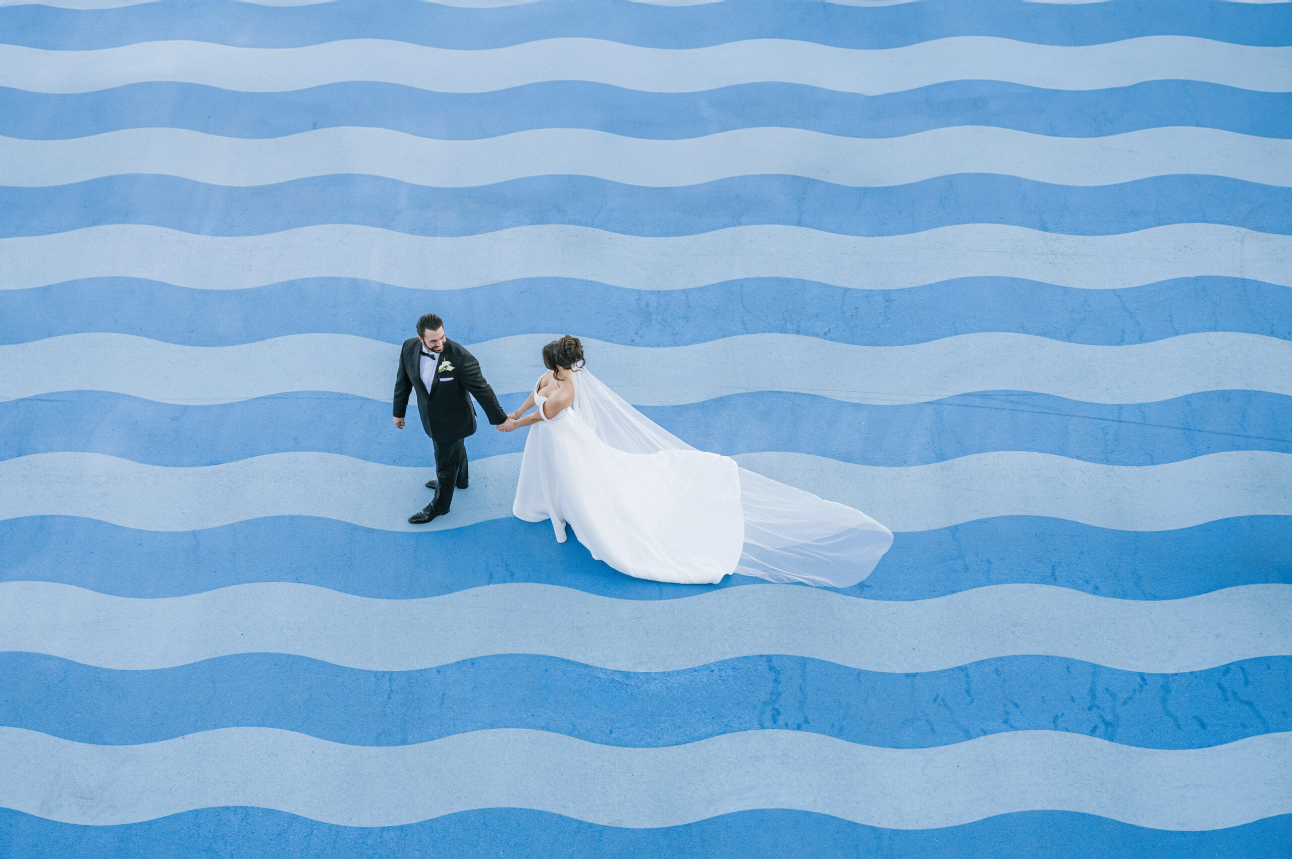 A bride in a flowing white gown walks hand in hand with her groom across a bold blue wave-patterned surface, creating a striking contrast and modern aesthetic.